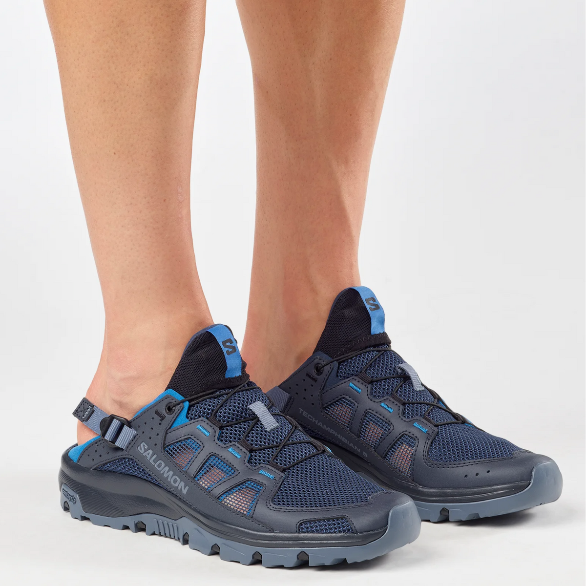 A pair of Salomon hiking shoes in navy blue and gray with bright blue accents, shown on a model's feet against a light gray studio background.