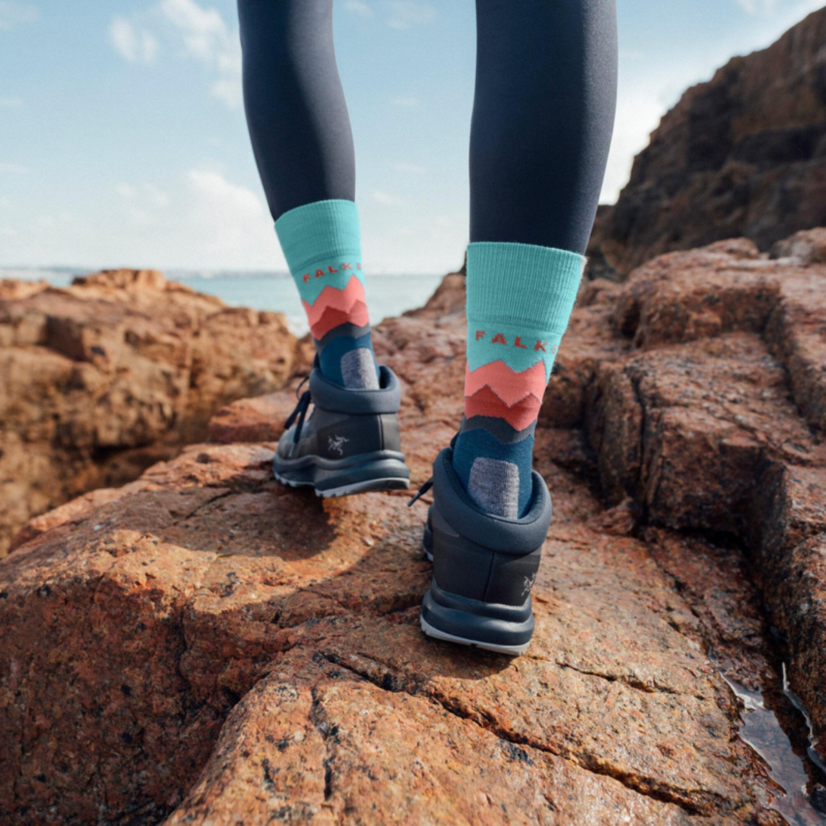 A person standing on rocky coastal ground wearing dark leggings, patterned socks, and dark athletic shoes.