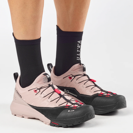 A person wearing black crew socks and Salomon Ultra hiking shoes in a dusty pink and black colorway, shown from the calves down against a light gray studio background.