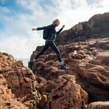 A woman in a black athletic jacket and leggings balances precariously on rugged brown rocks outdoors under a blue sky with clouds.