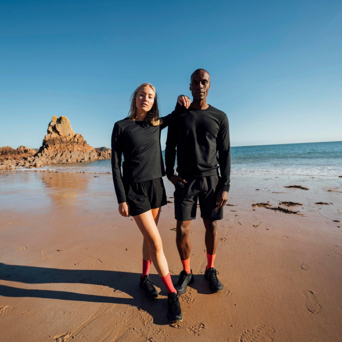 Two people standing on a sandy beach wearing matching black athletic long-sleeve tops and shorts, with the ocean, rocks, and blue sky in the background.