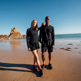 Two people standing on a sandy beach wearing matching black athletic long-sleeve tops and shorts, with the ocean, rocks, and blue sky in the background.