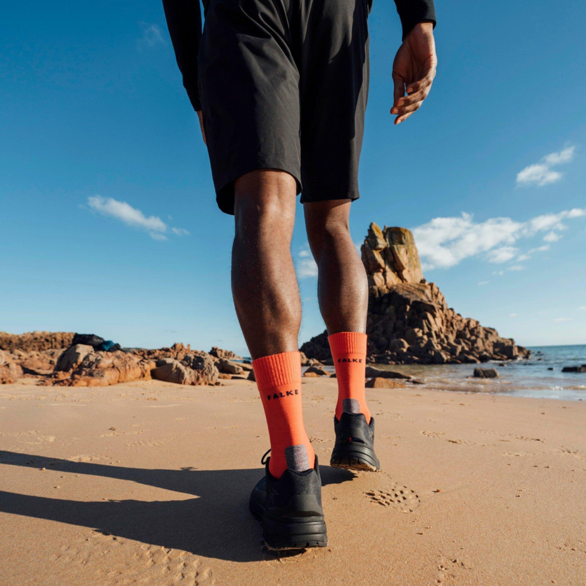 A person walking on a sandy beach wearing black athletic shorts, black running shoes, and bright orange crew socks with black branding.