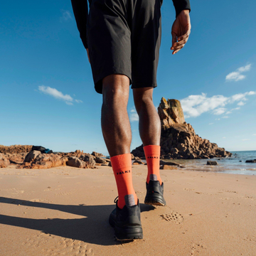 A person walking on a sandy beach wearing black athletic shorts, black running shoes, and bright orange crew socks with black branding.