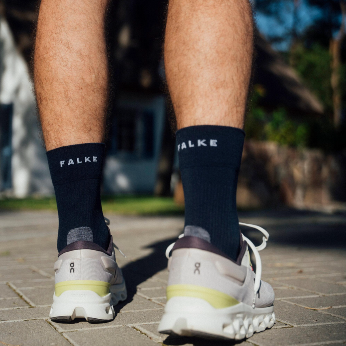 A person standing outdoors on a paved surface, shown from the back, wearing dark navy crew socks with the brand name 'FALKE' in white text and white athletic sneakers.