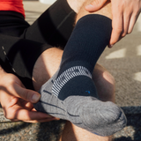 A close-up of a person sitting outdoors wearing a dark navy and heather gray athletic sock on their foot, with their hands adjusting the sock.
